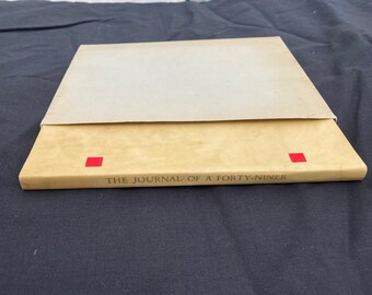 May include: A tan hardcover book titled The Journal of a Forty-Niner with two small red square accents on the spine, partially enclosed in a light beige paper slipcase. The book is positioned horizontally, displaying the title text clearly.
