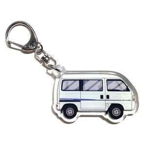 May include: White van keychain with a silver key ring. The van has blue stripes and is on a clear acrylic background.