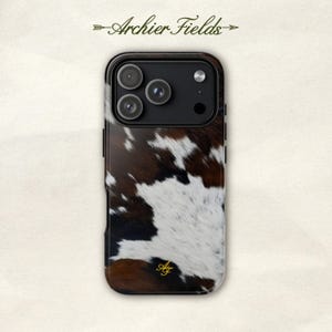 Op de afbeelding: Een rechthoekig smartphonehoesje met een bruin, zwart en wit koeienhuidpatroon. Het hoesje heeft een zwarte cameramodule met drie ronde lensopeningen en een flitser. De gouden monogramletters AF zijn onderaan op het hoesje gedrukt.