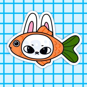 May include: A sticker featuring a cartoon fish with an orange body and a green tail fin. The fish has a white face with large black eyes and bunny ears. The background is a light blue grid pattern.