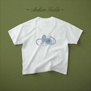 Może przedstawiać: Biały t-shirt z krótkim rękawem z niebieską grafiką przedstawiającą otwarty zegarek kieszonkowy z łańcuszkiem. Napis A Matter of Time jest wydrukowany na niebiesko pod zegarkiem. Nazwa marki Archier Fields znajduje się w kolorze ciemnozielonym.