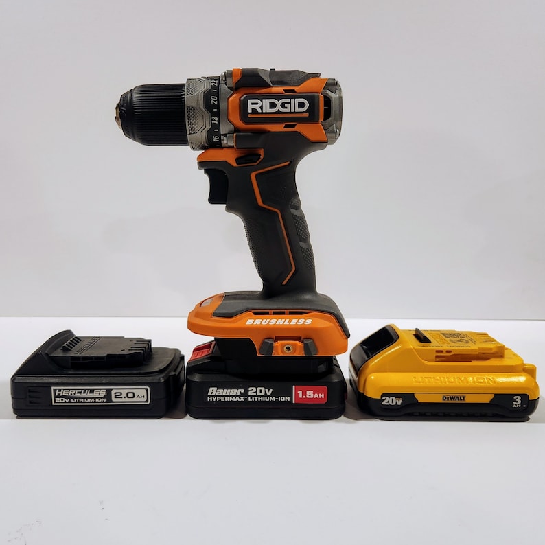 May include: A black and orange cordless drill with a brushless motor and a 20V battery. The drill is sitting on top of three different batteries: a black Hercules 20V lithium-ion battery, a black and orange Bauer 20V Hypermax lithium-ion battery, and a yellow Dewalt 20V lithium-ion battery.