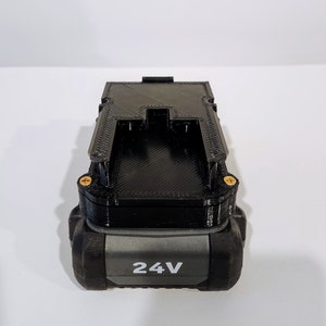Puede incluir: Una bater&iacute;a negra y gris con una etiqueta de 24V. La bater&iacute;a tiene una forma rectangular con una superficie texturizada y dos tornillos dorados en el lateral.