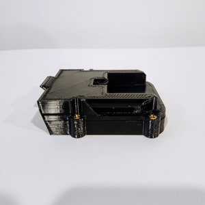 Puede incluir: Una caja negra impresa en 3D con tornillos dorados. La caja tiene forma rectangular y una peque&ntilde;a abertura en la parte superior.