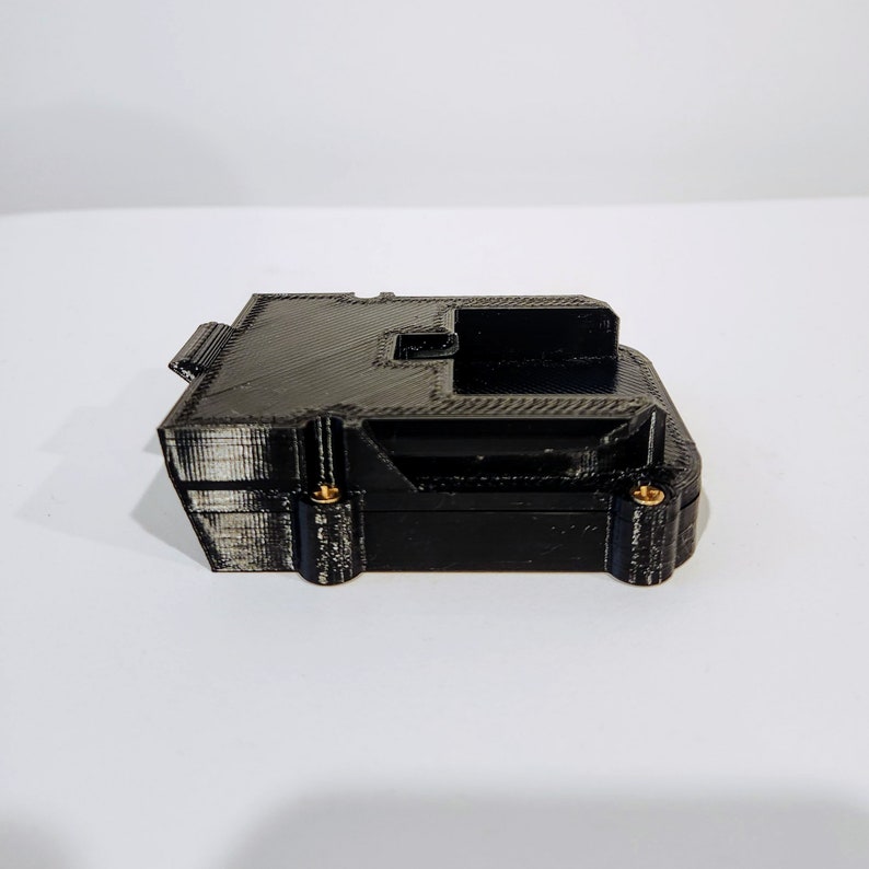 Puede incluir: Una caja negra impresa en 3D con tornillos dorados. La caja tiene forma rectangular y una peque&ntilde;a abertura en la parte superior.