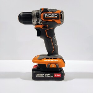 May include: An orange and black cordless drill with a black handle and a battery pack that says "Bauer 20V HYPERMAX Lithium-Ion 1.5Ah". The drill has a black chuck and a silver band with markings. The drill is labeled "RIDGID" in orange letters.
