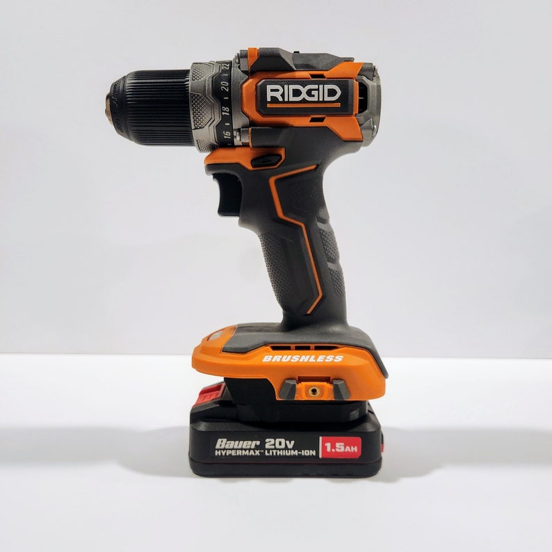 May include: An orange and black cordless drill with a black handle and a battery pack that says "Bauer 20V HYPERMAX Lithium-Ion 1.5Ah". The drill has a black chuck and a silver band with markings. The drill is labeled "RIDGID" in orange letters.