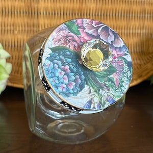 May include: A glass jar with a floral patterned lid and a clear crystal knob. The lid has a black and white polka dot trim.