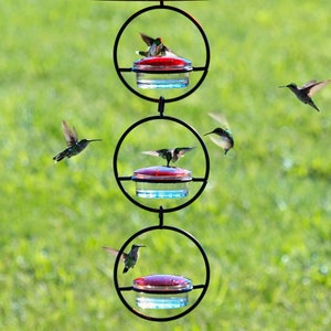 Three Hummingbird feeders hang in a straight line like a chain. Each feeder includes a vertical circular metal outer band. At the bottom of each circle, a clear easy to wash dish is displayed with a removable red glass lid.