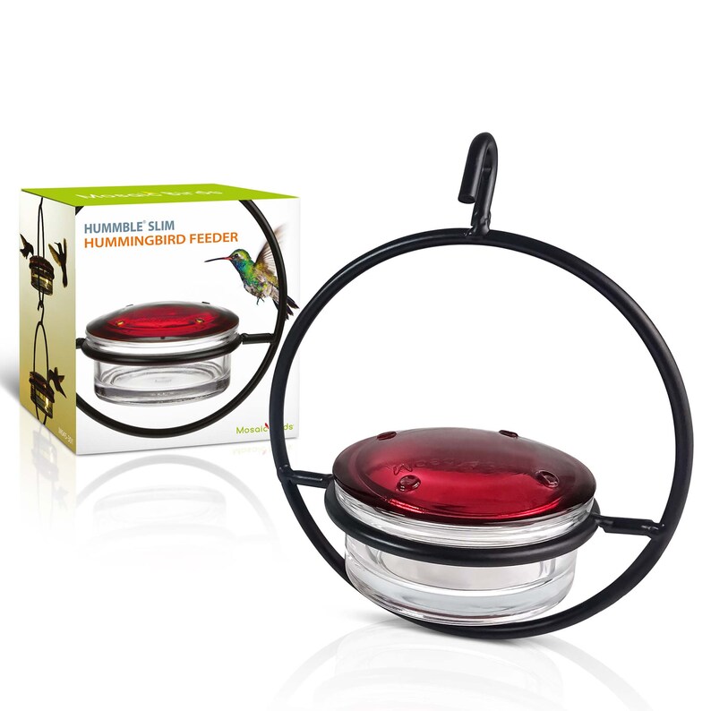 May include: A black metal hummingbird feeder with a clear glass reservoir and a red lid. The feeder is suspended by a curved hook and has a circular frame. The box next to it reads "HUMMBLE SLIM HUMMINGBIRD FEEDER".