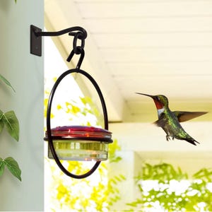 May include: A black metal hummingbird feeder hanging from a wall bracket. The feeder has a glass jar with a red top and a clear base. A hummingbird with green and red feathers is flying nearby.