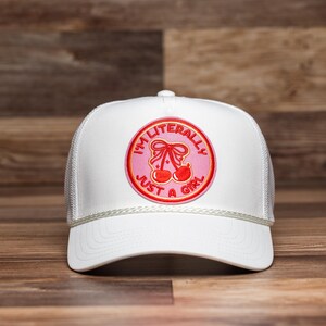 Op de afbeelding: Witte truckerpet met een mesh achterkant en een gebogen klep. De pet heeft een cirkelvormige patch met de tekst "I'M LITERALLY JUST A GIRL" rond een kersendesign in roze en rood.