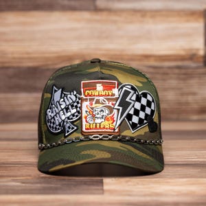 Op de afbeelding: Een camouflage trucker cap met een kettingdetail. De pet heeft verschillende patches, waaronder een schedel met de tekst "Cowboy Killers", een bliksem en een schoppen met de tekst "Raising Hell".