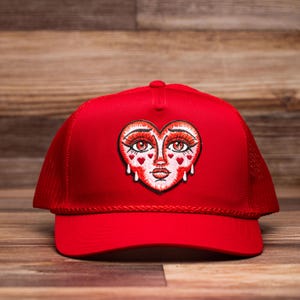 May include: A vibrant red trucker cap with a heart-shaped face patch. The patch features red eyes, lips, and teardrops, with detailed embroidery. The cap has a mesh back and a curved peak.