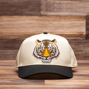May include: A beige baseball cap with a black brim and a large embroidered tiger face on the front.
