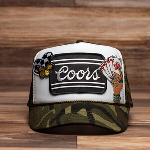 May include: A trucker hat with a camouflage brim and a white front panel. The front features a black patch with the word "Coors" in white script, a butterfly, and a hand holding playing cards. The hat has a mesh back.