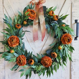 May include: A burnt orange and green floral wreath with a brown satin ribbon bow. The wreath is made of silk flowers and greenery.