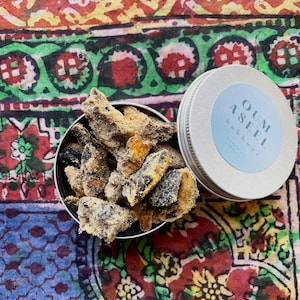 May include: A small, open, silver tin filled with pieces of dark brown and tan bukhoor, with a matching lid next to it. The lid has the words "OUM ASEEL" and "bukhoor" printed on it. The background is a colorful patterned fabric.