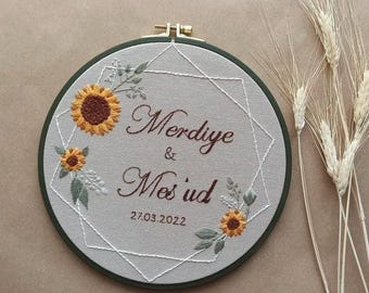 May include: Embroidered hoop art with a geometric design, featuring two sunflowers, green leaves, and the names "Merdiye & Mes'ud" in brown cursive lettering. The date "27.03.2022" is also embroidered below the names.