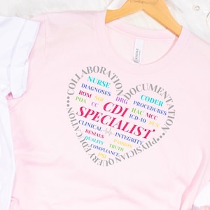 May include: Two t-shirts, one white and one pink, with a circle design featuring the words "CDI Specialist" surrounded by other words related to healthcare documentation, such as "nurse", "coder", "diagnoses", "procedures", "integrity", and "compliance".
