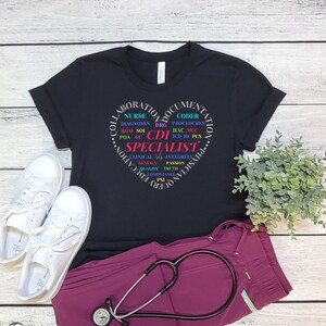 May include: Black t-shirt with a heart-shaped design featuring the text "Collaboration, Documentation, Physician Education" and "CDI Specialist" in a colorful font. The text includes abbreviations related to healthcare, such as "DRG", "ICD-10", and "POA".