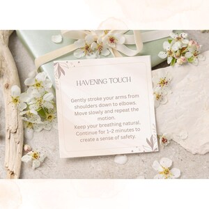 May include: A square card with the text "HAVENING TOUCH" and instructions for a self-soothing technique. The card is surrounded by white flowers and rests on a light-colored surface, with a wrapped gift in the background.