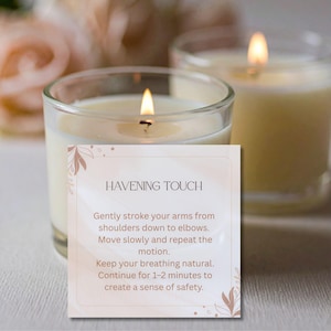 May include: Two lit candles in clear glass holders. A card in front of the candles reads "HAVENING TOUCH" with instructions for a self-soothing technique. The candles have a soft, warm glow, suggesting relaxation and comfort.