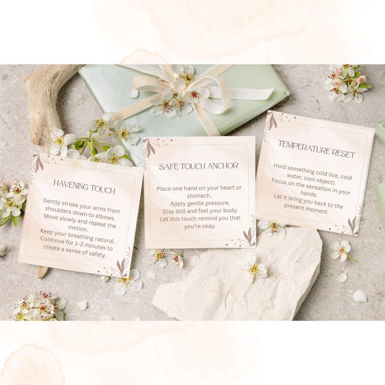 May include: Three cards with text instructions for self-soothing techniques, including "Havening Touch," "Safe Touch Anchor," and "Temperature Reset." The cards are surrounded by white flowers and a wrapped gift with a ribbon.