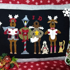 Może przedstawiać: Dekoracyjna poduszka świąteczna z czarnym tłem z aplikacjami reniferów śpiewających kolędy. Renifery mają na sobie szaliki i płaszcze, a u ich stóp znajdują się pies i kot. Choinka i płatki śniegu dopełniają świąteczny design.
