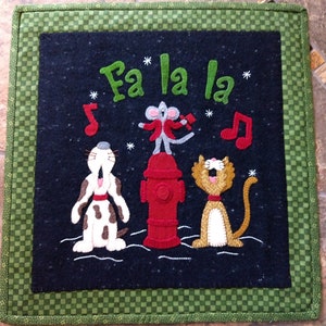 May include: A felt Christmas wall hanging with a black background and a green border. The text "Fa la la" is embroidered in green. A red fire hydrant is in the center with a white mouse, a brown and white dog, and a white and orange cat singing around it.