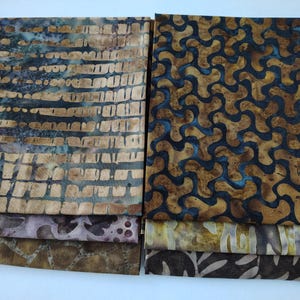 May include: Two stacks of folded fabric featuring batik patterns. The left stack displays horizontal brown and tan stripes, a purple mottled design, and a brown crackle print. The right stack shows a dark blue and tan wave pattern, a yellow mottled print, and a dark brown leaf motif.