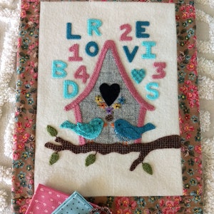 Könnte beinhalten: Ein Wandbehang aus Filz mit einem rosa und grauen Vogelhaus, auf dem zwei blaue Vögel auf einem Ast sitzen. Das Vogelhaus hat ein schwarzes Herz und die Buchstaben "LOVE" sind in blauen und rosa Filzbuchstaben um das Vogelhaus herum geschrieben.