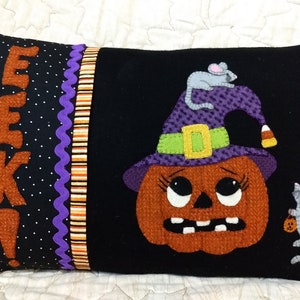 May include: A black pillow with a purple, orange, and white striped border. The pillow features a felt applique of a pumpkin wearing a witch's hat with a mouse on top. The pumpkin has a surprised expression and the text "EEK!" is on the left side of the pillow.