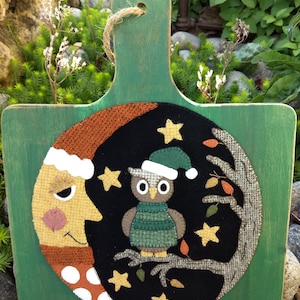 May include: A green wooden cutting board with a felt appliqué of a moon, owl, and stars. The moon is wearing a Santa hat and has a white polka dot scarf. The owl is also wearing a Santa hat and is perched on a branch. The background is black with gold stars.