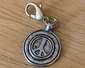 Könnte beinhalten: Ein silberfarbener Peace-Zeichen-Anhänger mit einem goldfarbenen Verschluss. Das Peace-Zeichen befindet sich in der Mitte eines kreisförmigen Anhängers. Der Anhänger ist zum Anbringen an einer Halskette, einem Armband oder einem Schlüsselanhänger gedacht.
