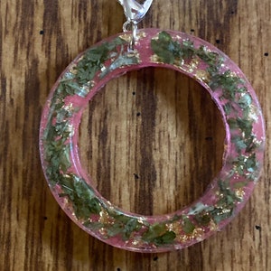 Könnte beinhalten: Ein rosafarbener Harzring mit eingebetteten goldenen Flocken und grünen Blättern. Der Ring hat einen silbernen Metallverschluss.