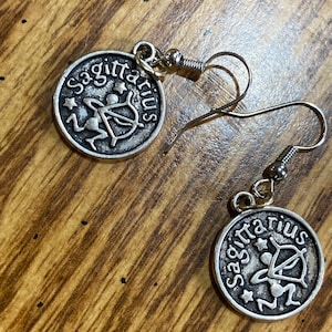 May include: A pair of silver Sagittarius earrings with a hook closure. The earrings feature a circular design with the zodiac symbol of Sagittarius, an archer with a bow and arrow, and the word "Sagittarius" written around the edge.