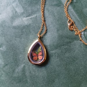May include: A gold chain necklace with a teardrop-shaped pendant. The pendant contains a preserved butterfly with orange wings and green foliage.