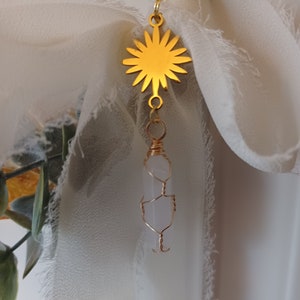 May include: A gold sun charm with a clear quartz crystal pendant hanging from it. The charm is attached to a white fabric ribbon.