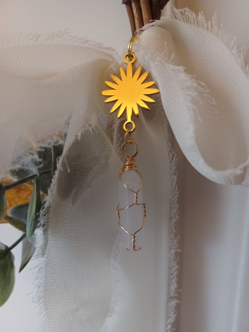 May include: A gold sun charm with a clear quartz crystal pendant hanging from it. The charm is attached to a white fabric ribbon.