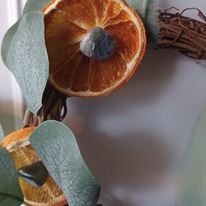 May include: A dried orange slice with a gray and green stone in the center, surrounded by green eucalyptus leaves on a brown twig wreath.