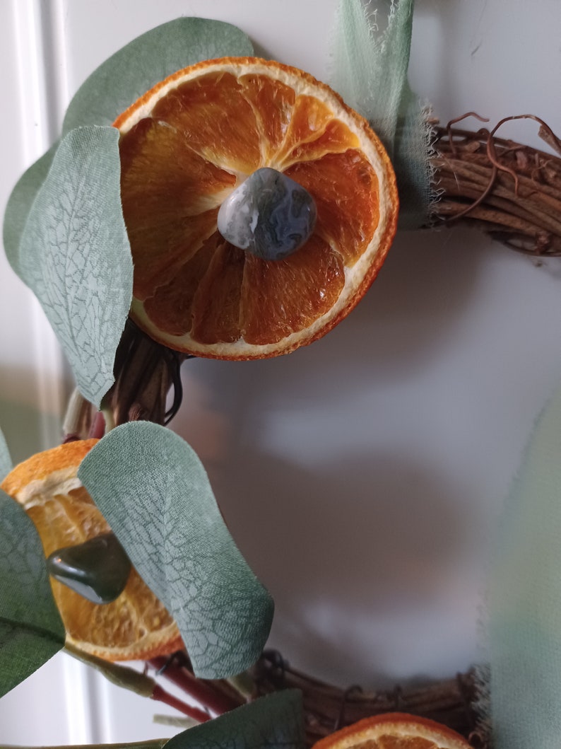 May include: A dried orange slice with a gray and green stone in the center, surrounded by green eucalyptus leaves on a brown twig wreath.