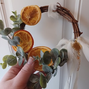 May include: A rustic twig wreath decorated with dried orange slices and eucalyptus leaves. A white fabric ribbon with a gold charm hangs from the wreath.