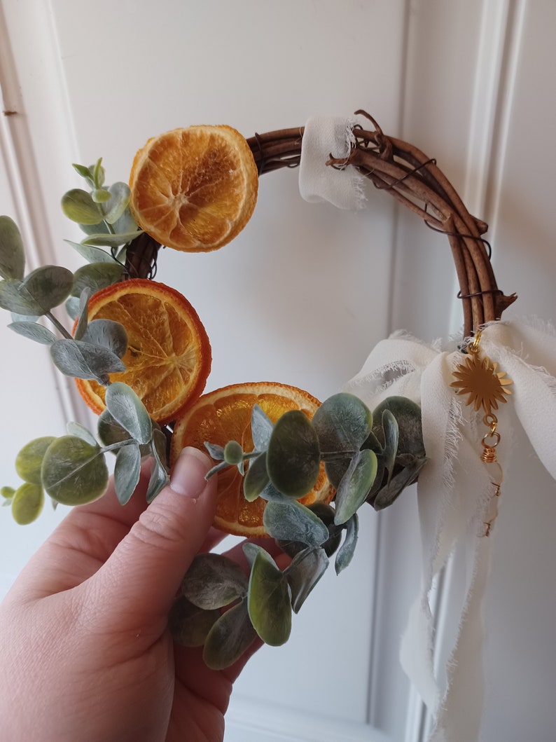 May include: A rustic twig wreath decorated with dried orange slices and eucalyptus leaves. A white fabric ribbon with a gold charm hangs from the wreath.