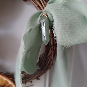 May include: A green crystal wrapped in gold wire hangs from a twig wreath with a sage green ribbon.