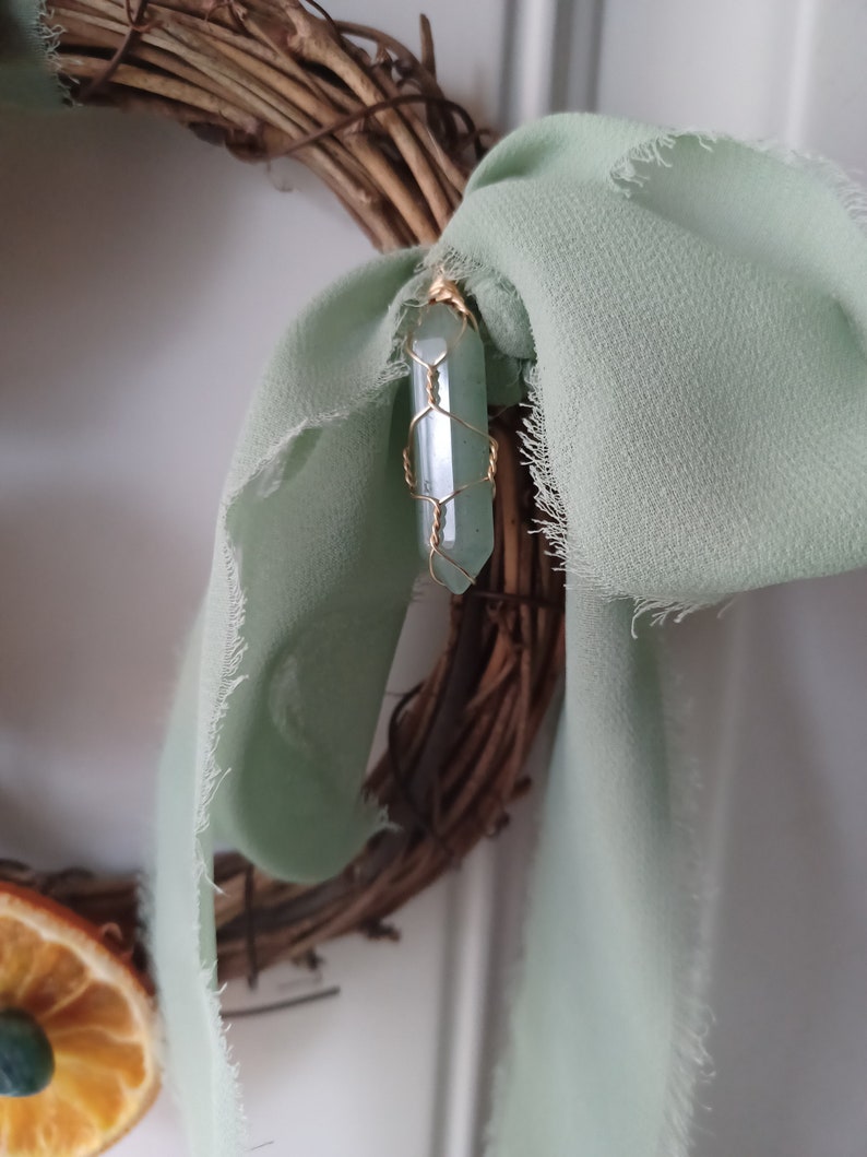 May include: A green crystal wrapped in gold wire hangs from a twig wreath with a sage green ribbon.
