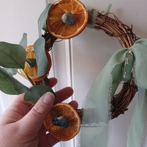 May include: A rustic twig wreath decorated with dried orange slices and green eucalyptus leaves. Each orange slice has a small green gemstone in the center. A sage green ribbon is tied around the wreath.