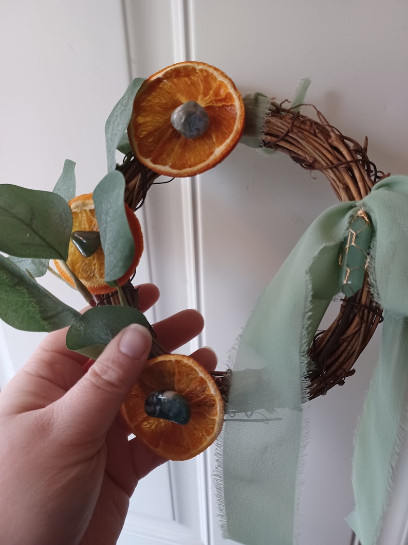 May include: A rustic twig wreath decorated with dried orange slices and green eucalyptus leaves. Each orange slice has a small green gemstone in the center. A sage green ribbon is tied around the wreath.