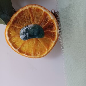 May include: A dried orange slice with a green and white agate stone on top. The orange slice is on a white surface.