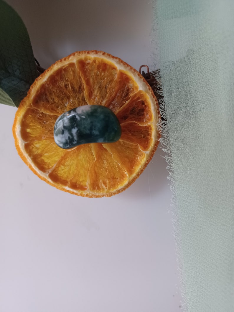 May include: A dried orange slice with a green and white agate stone on top. The orange slice is on a white surface.